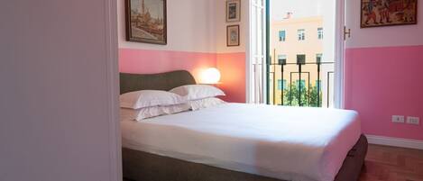 A bedroom with a bed, a nightstand, a window with a view of buildings, and a door leading to another room.