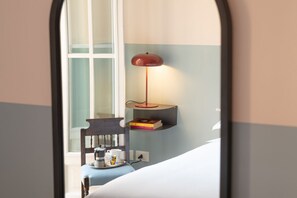 Una habitación con cama, una silla, una mesita con lámpara y una estantería con libros.