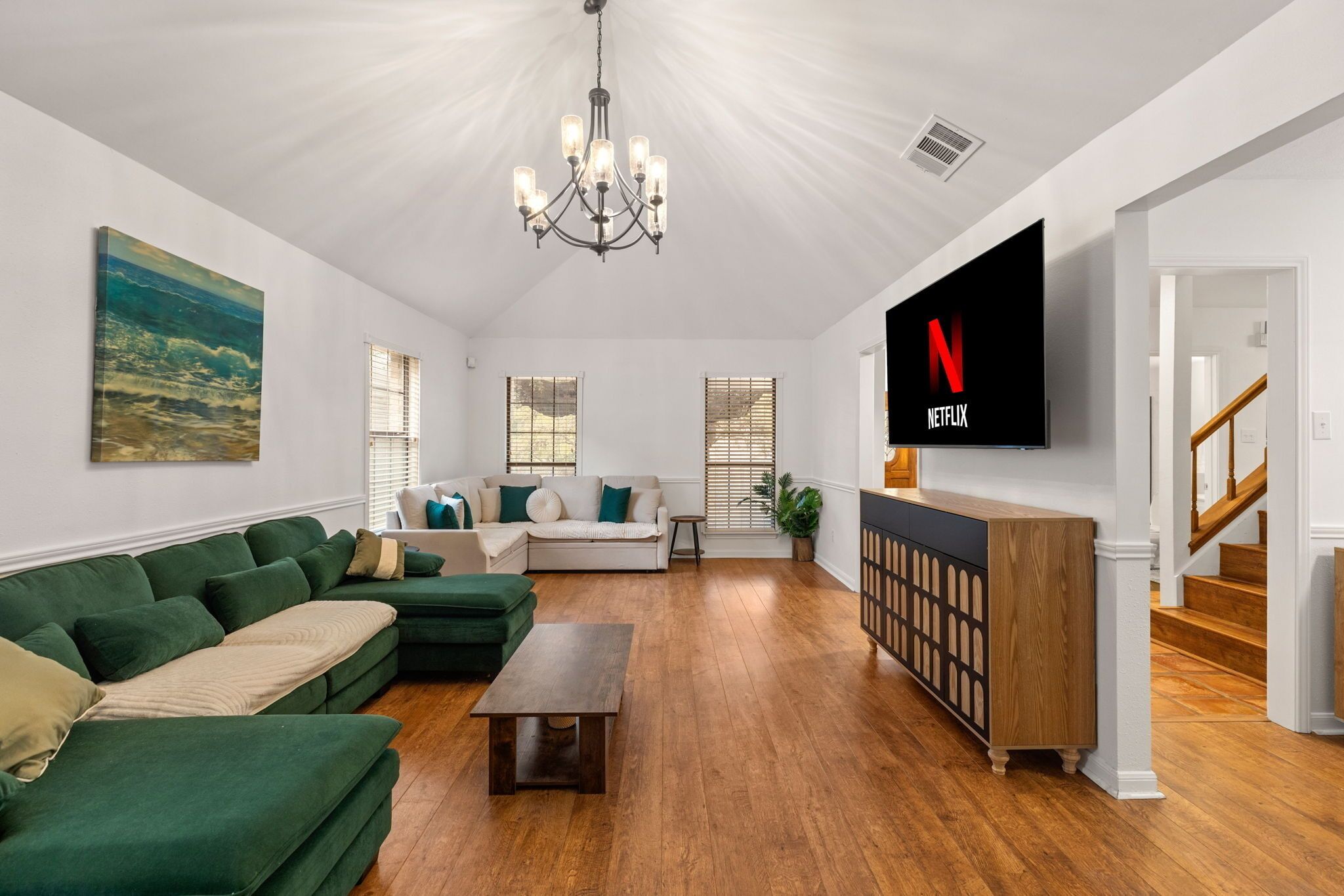 A living room with a green sofa, a white sofa, a wooden coffee table, a TV on the wall, and a staircase.