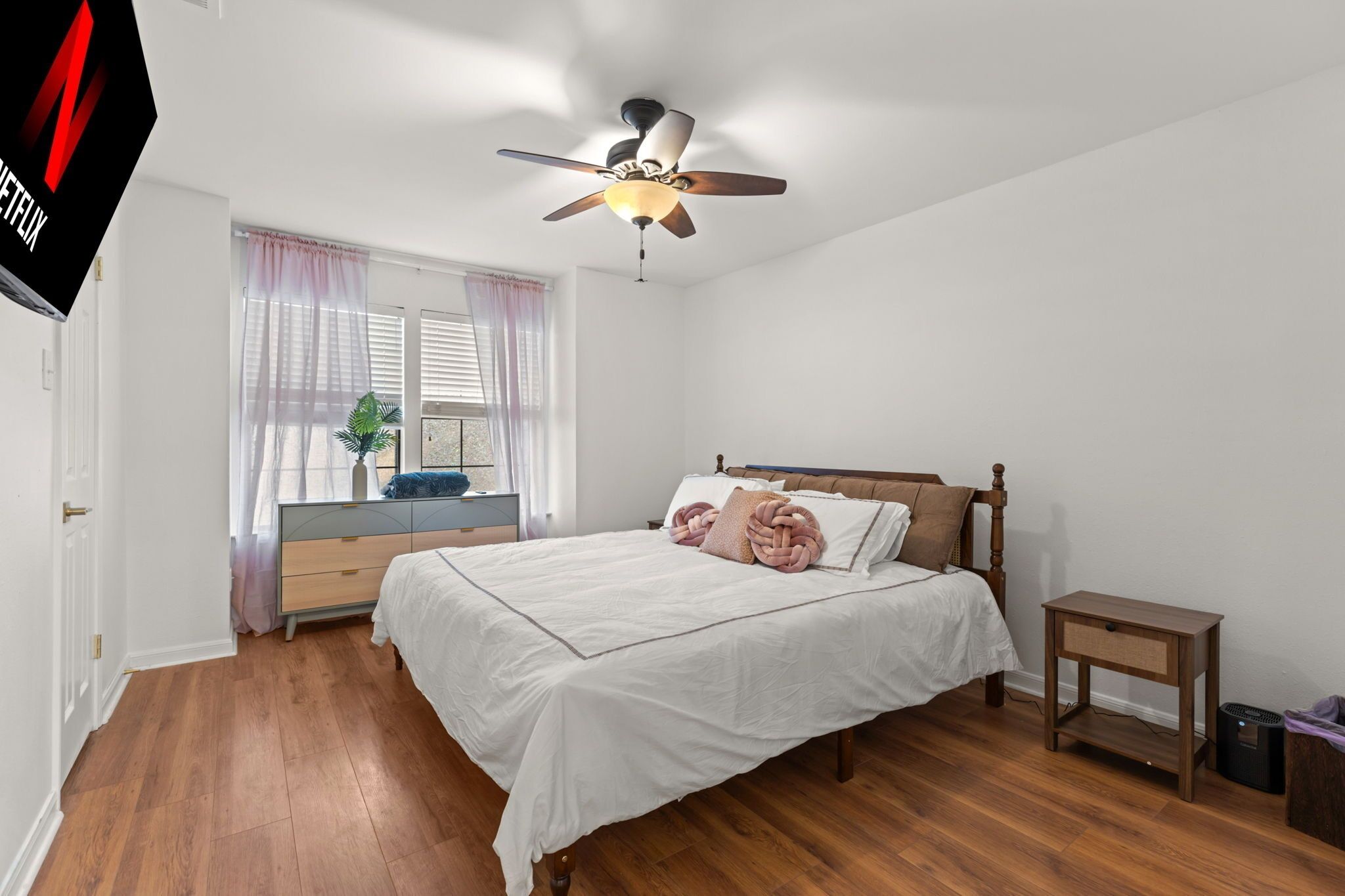 A bedroom with a bed, a ceiling fan, a bedside table, a dresser, and a window with curtains.