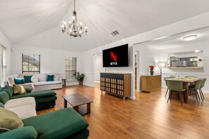 Ein modernes Wohnzimmer mit Flachbildfernseher, grünen Sofas, Holzböden und einem Essbereich mit Tisch und Stühlen.
