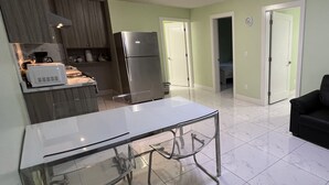 Cozinha moderna com frigorífico, micro-ondas e máquina de café. Há uma mesa de jantar com cadeiras e um sofá preto na área de estar.