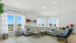 A modern living room with a sectional sofa, a blue armchair, a small table, and a large window offering a view of the outdoors. - Grenadine Dreams (Gulf Shores)