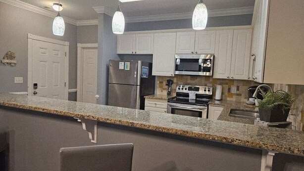 A kitchen with granite countertops, white cabinets, a stainless steel refrigerator, a microwave, and an oven. There are two pendant lights, a potted plant, and a welcome sign on the wall. - Sea Bound | Golf Getaway + Pool | Myrtle Beach (Myrtle Beach)