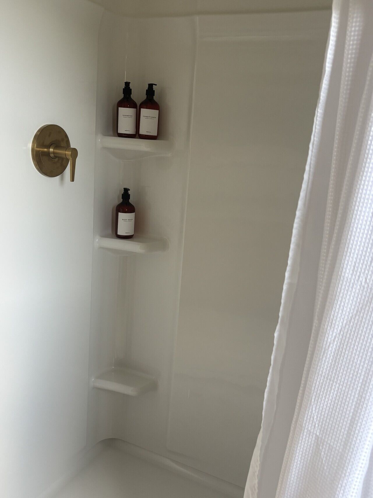 A bathroom with a shower, a brass door handle, and three shelves holding bottles.