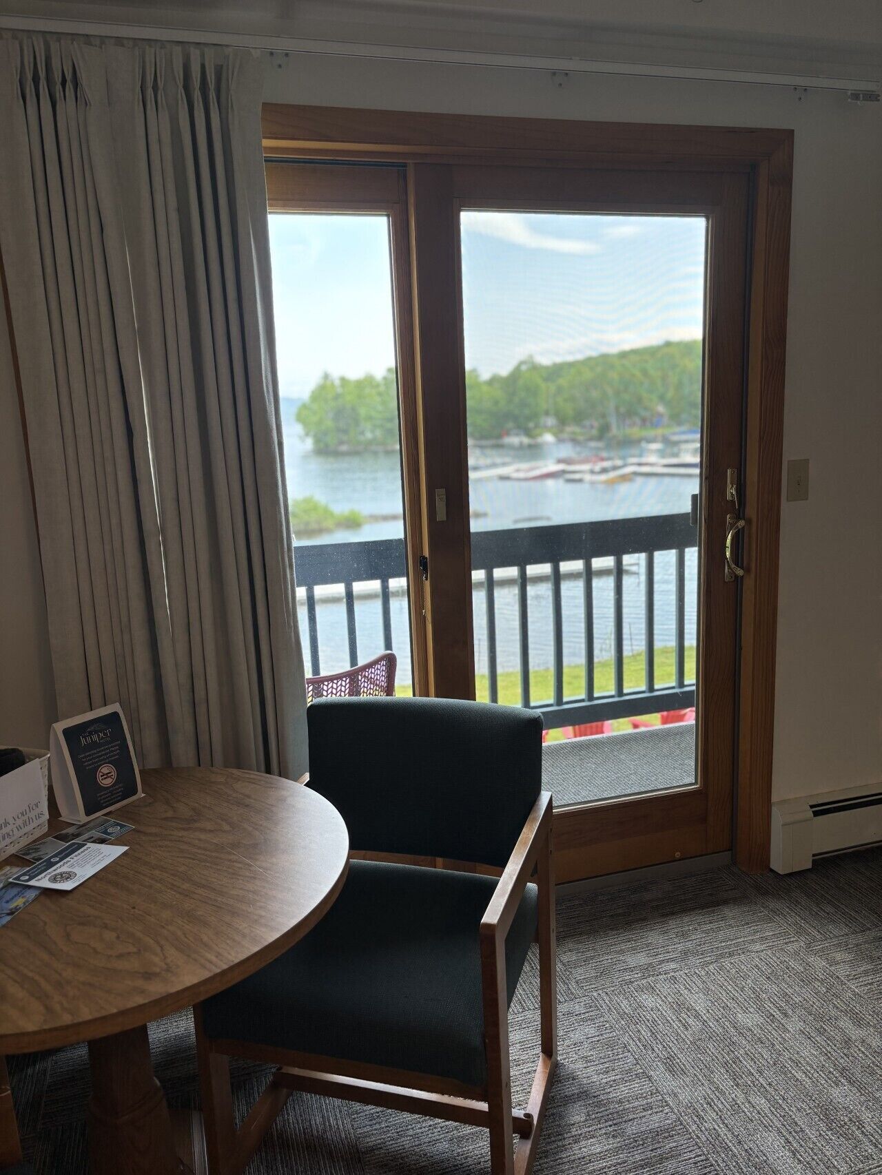 A room with a wooden table, a chair, a view of a lake, and a balcony with a railing.