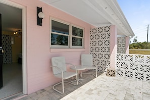 Un edificio rosa con dos sillas blancas y una mesa en el exterior, una valla con diseño y una puerta que da al interior.