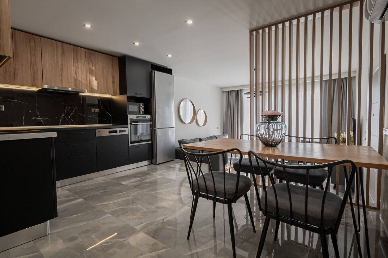 A modern kitchen with dark cabinetry, a wooden dining table, and a large window with vertical blinds.