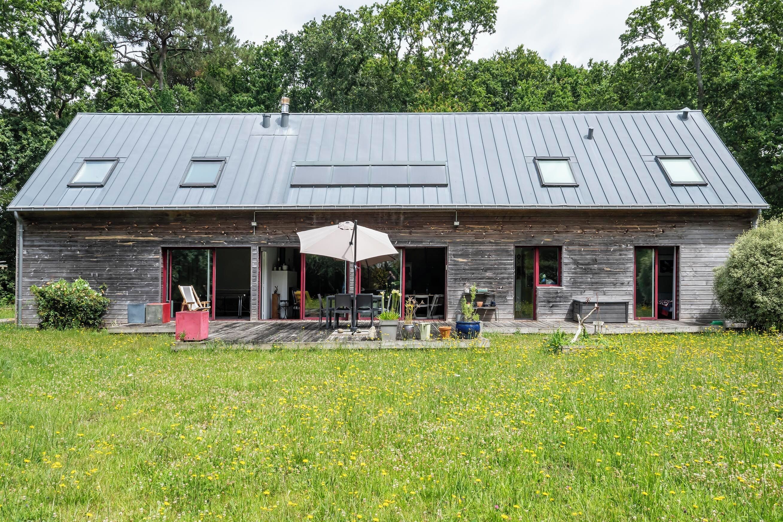 Een modern houten huis met een ruime open patio, een overdekte zithoek en een veld met gele bloemen op de voorgrond.
