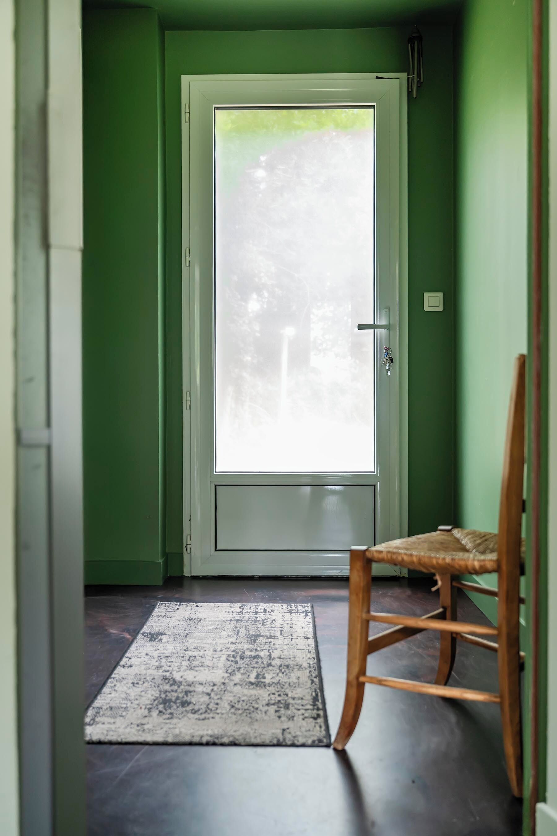 Una habitación con pared verde, una silla de madera, un tapete y una puerta con un ventanal de vidrio esmerilado.