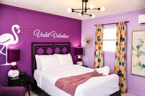 Ein Schlafzimmer mit einem Bett, einem Nachttisch, einer Lampe, einer Wand mit Flamingo-Dekoration und einem Fenster mit Vorhängen.