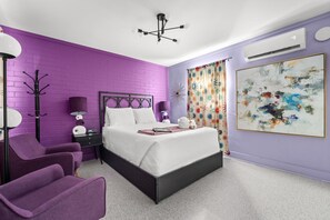 A bedroom with a purple brick wall, a bed with white bedding, a black headboard, a nightstand with a lamp, a purple chair, and a large abstract painting. - Violet Valentino | Purple Boutique Retreat (Port Saint Joe)