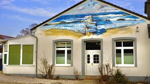 A building with a mural of a lighthouse and waves, a white door, and two windows with shutters. - Country house Wendy 1 semi-detached house 250 m² 6 bedrooms, 4 bathrooms, garden (Gägelow)