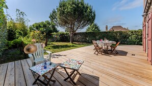 Une terrasse en bois avec des meubles d’extérieur, un grand arbre et une haie en arrière-plan.