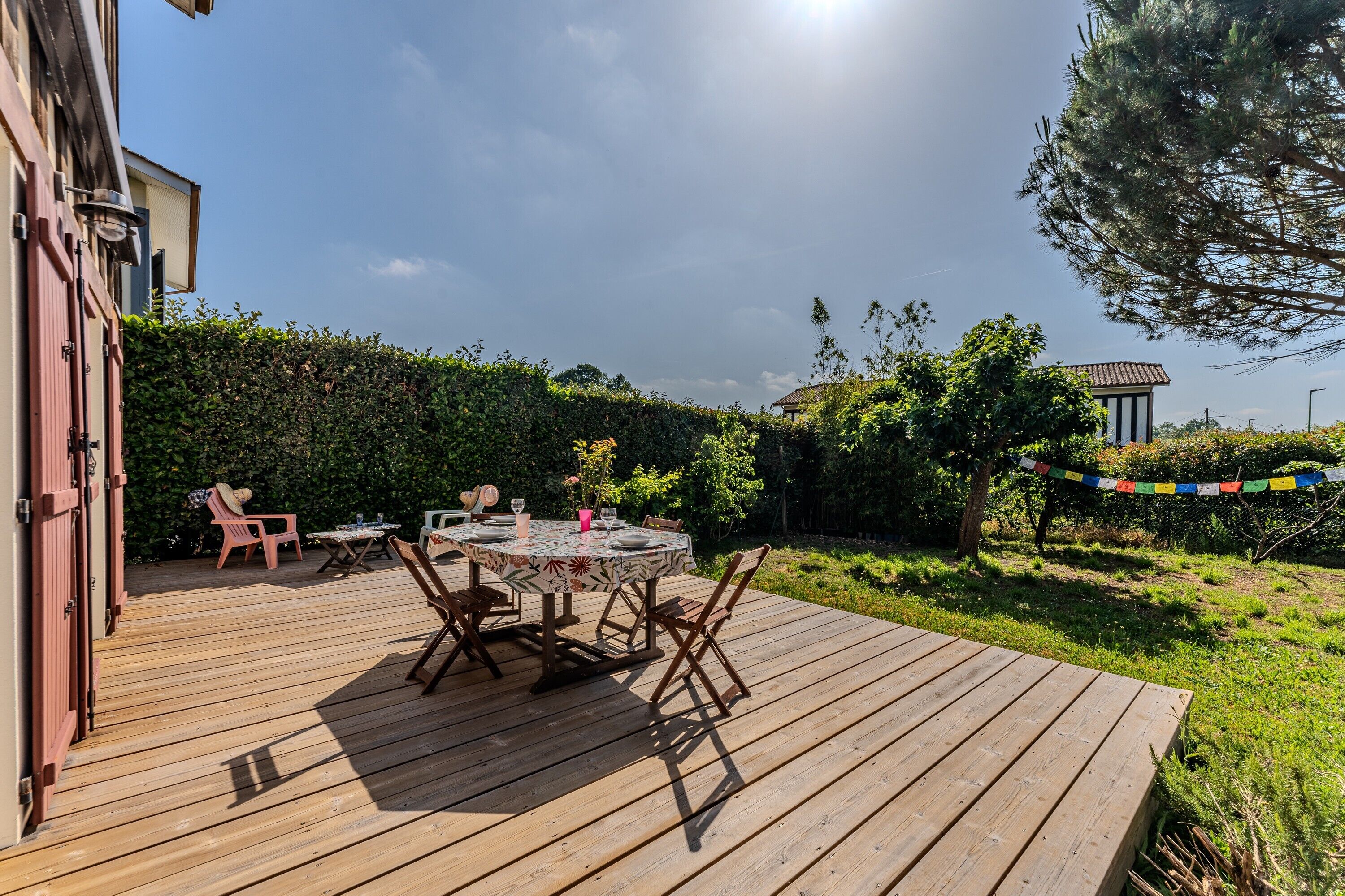 Une terrasse en bois avec une table ronde et des chaises, entourée de verdure et sous un ciel dégagé.