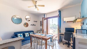 Een kamer met een blauwe bank, een houten eettafel met stoelen, een plafondventilator en uitzicht door een glazen deur.