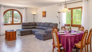 A living room with a sectional sofa, a dining table with chairs, and a large window with a view of greenery. - Villa Papillon by Villa Plus (Calp)
