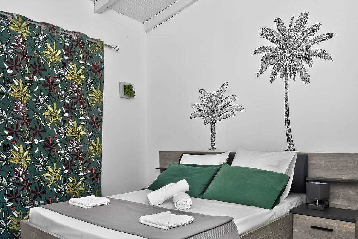 Una cama bien tendida con ropa de cama verde y blanca, una mesita de noche con una lámpara y una pared decorada con dos dibujos de palmeras.