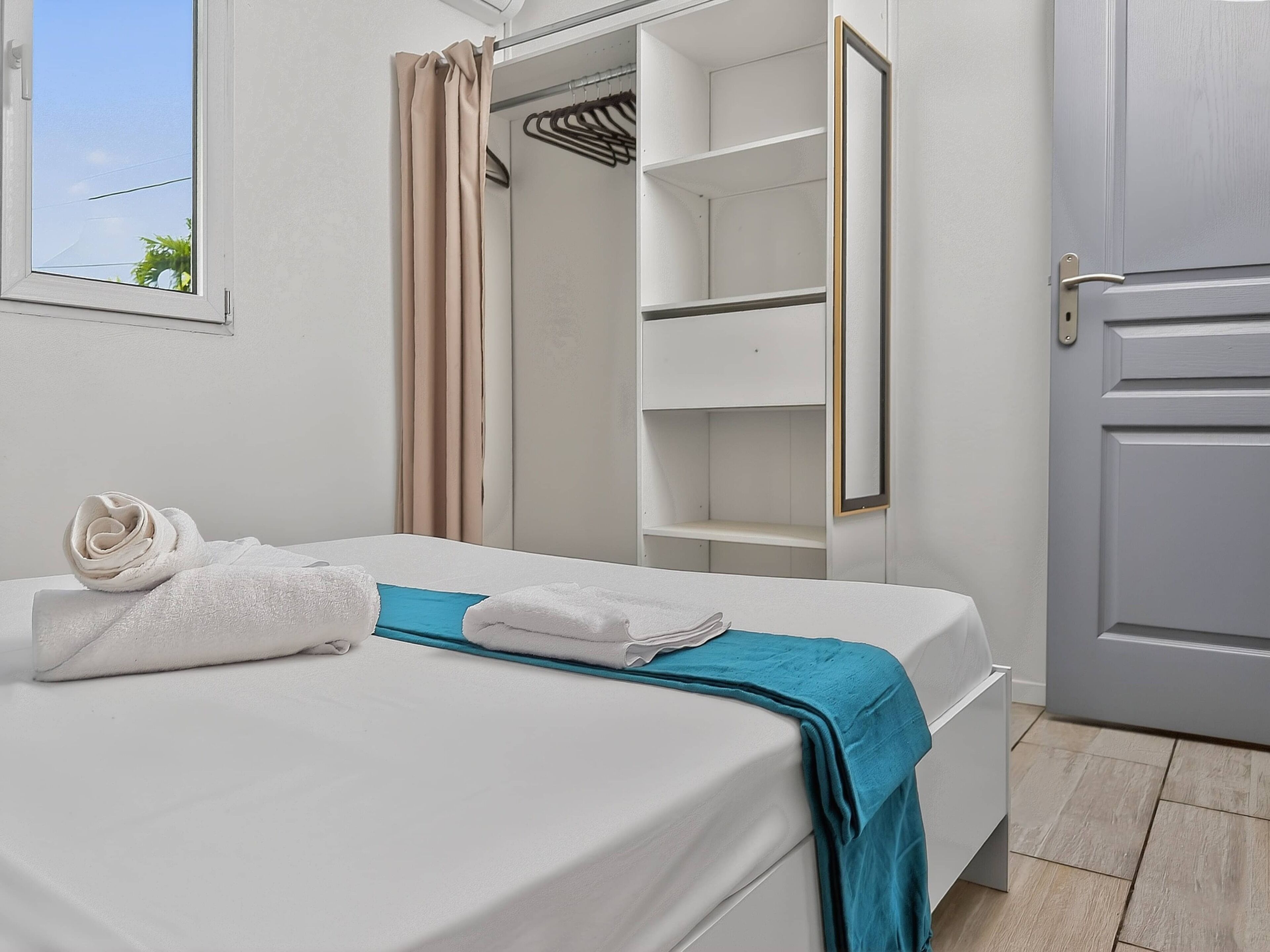 Un lit bien fait, avec des serviettes à portée de main, une armoire dont les portes sont ouvertes et un miroir.