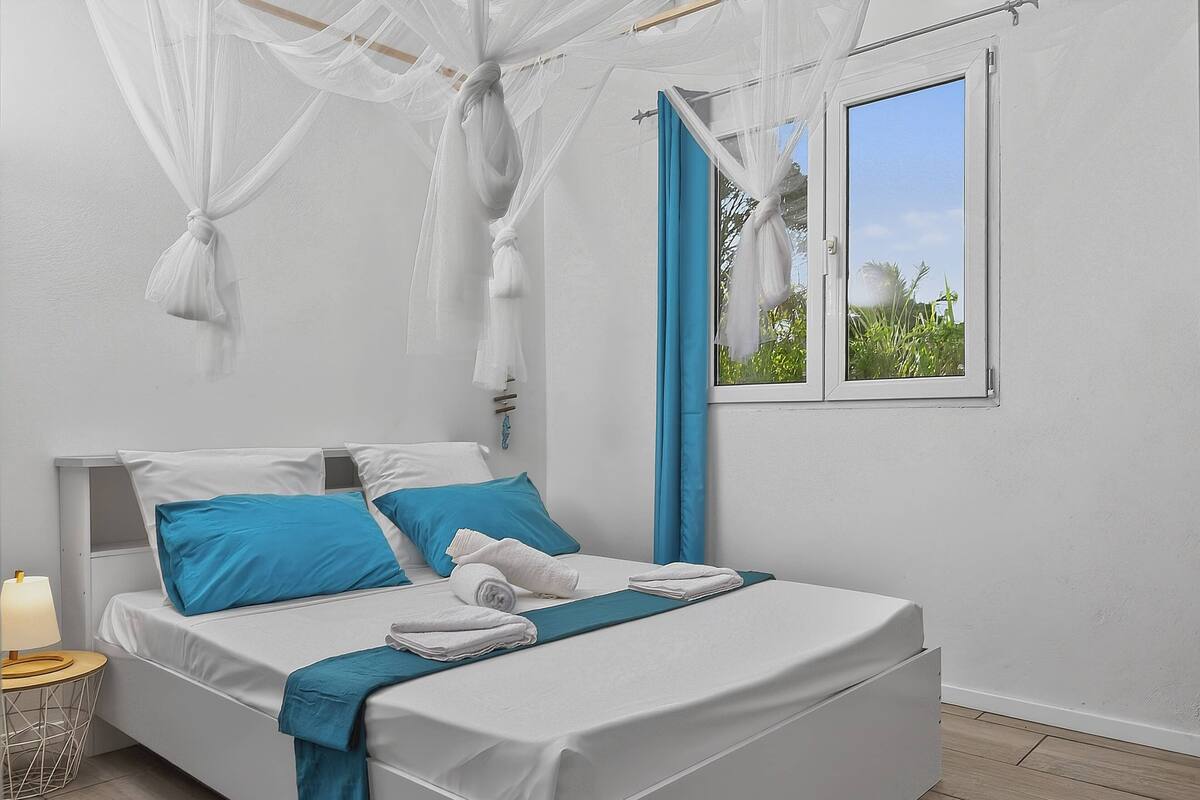 Una cama con ropa de cama blanca y azul, una mesita de noche con una lámpara y una ventana con vistas a zonas verdes.