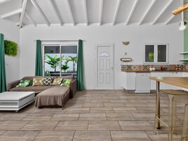 A modern living space with a sofa, a dining table, a kitchen area, and a door leading to a balcony with plants.