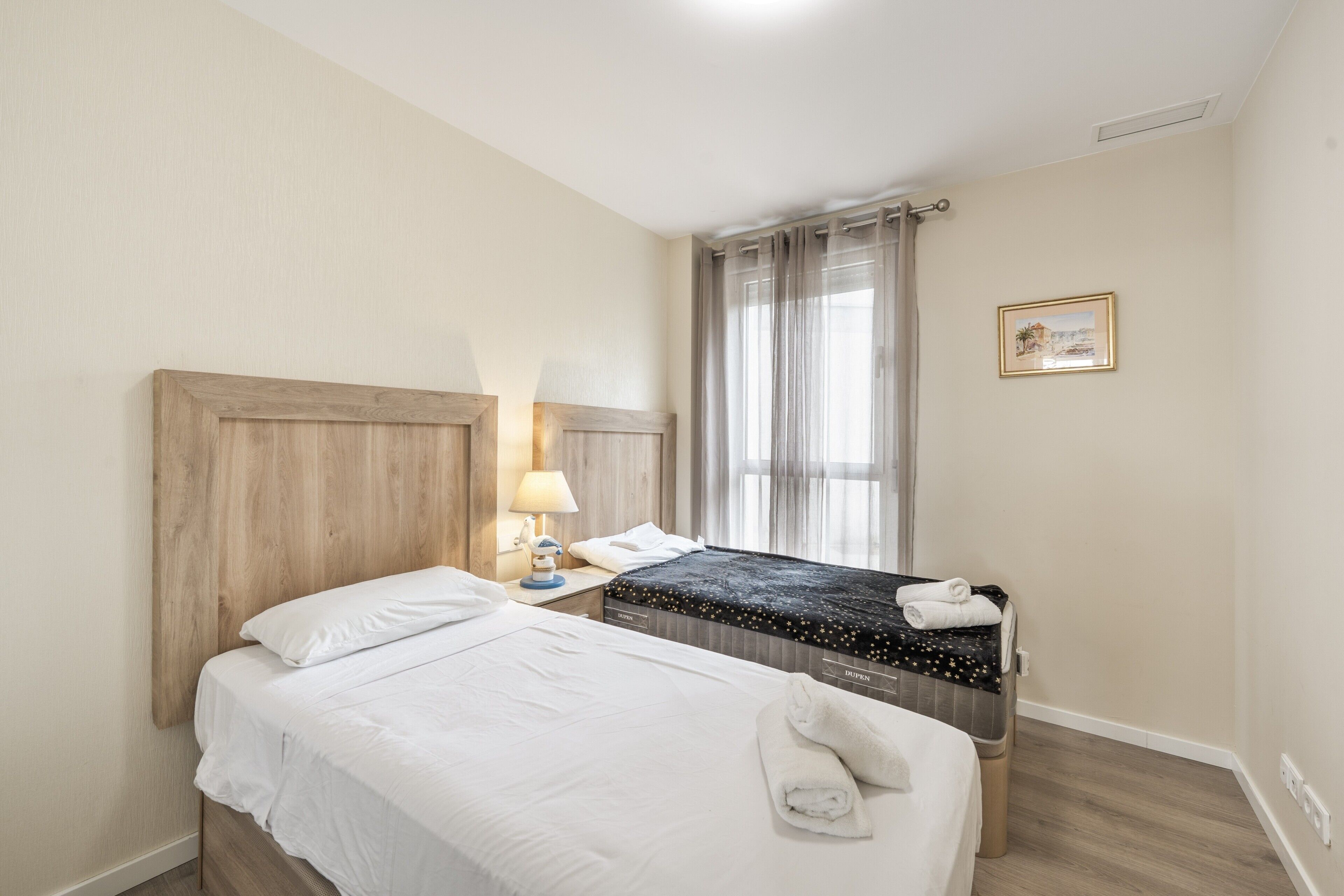 A hotel room with two beds, a nightstand with a lamp, a window with curtains, and a framed picture on the wall.