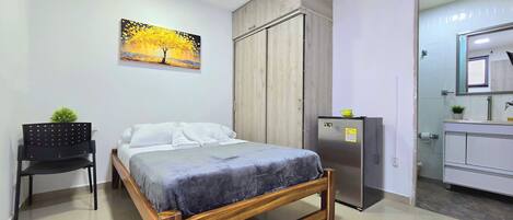 A bedroom with a bed, a chair, a wardrobe, a refrigerator, and a bathroom with a mirror.