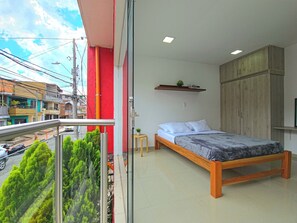 A bedroom with a bed, a small table, a shelf with books, and a view of the street. - HOTEL CASA MEGUA3 (Medellín)