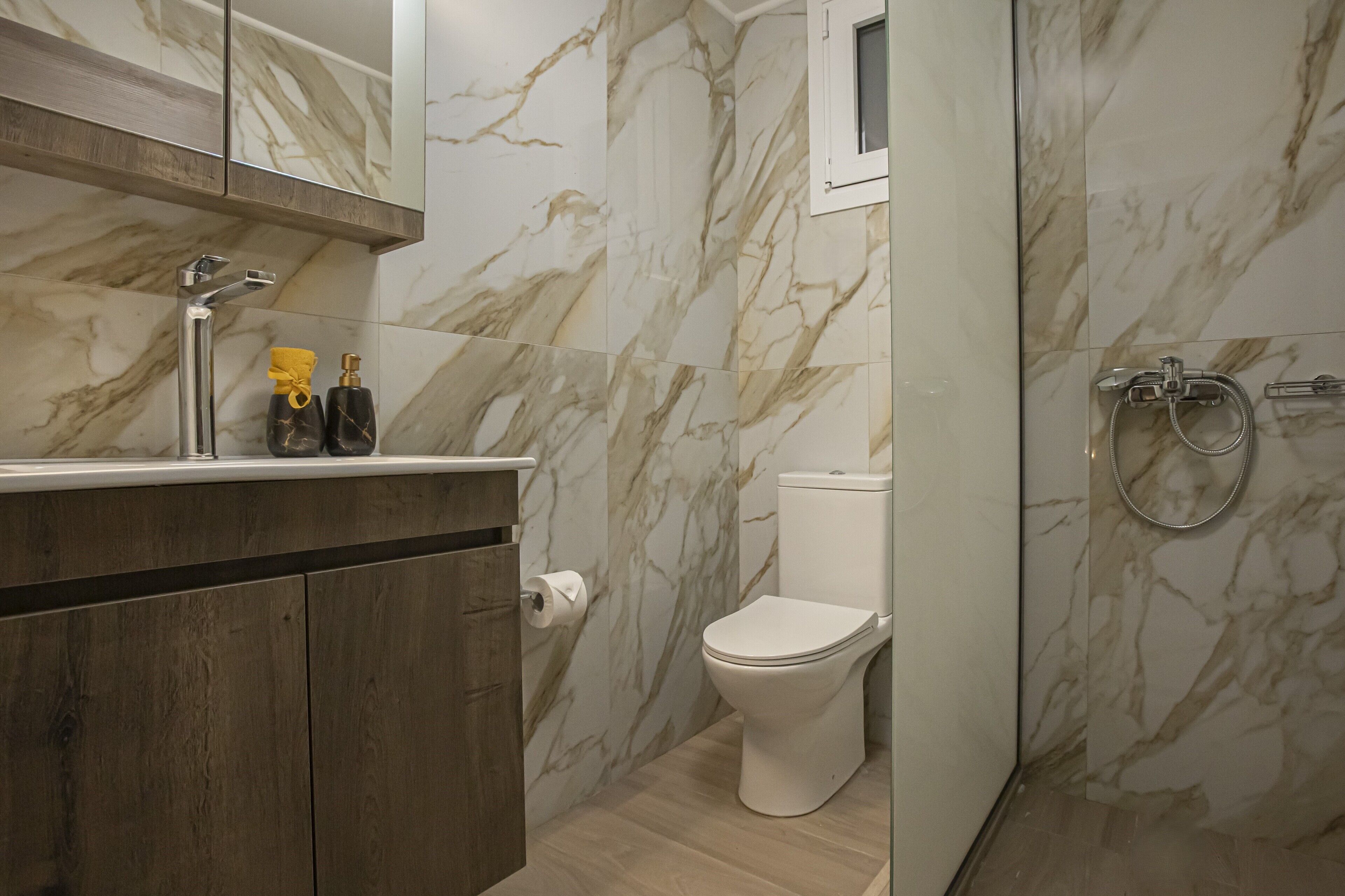 Un baño con paredes de mármol, un inodoro blanco, una ducha con cabezal de ducha manual y un tocador de madera con espejo.