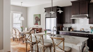A modern kitchen with dark cabinets, a central island with bar stools, pendant lighting, and a large window. - SoCo Craftsman ~ Walk to Shops, Cafés, Parks (Austin)