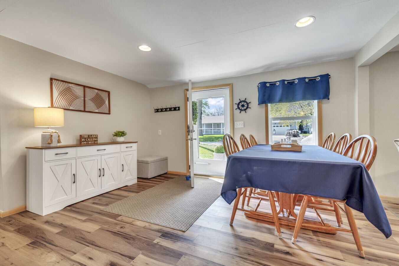 Uma sala de jantar com uma mesa posta para quatro pessoas, um armário branco com gavetas, um tapete no chão e uma vista pela porta para o exterior.