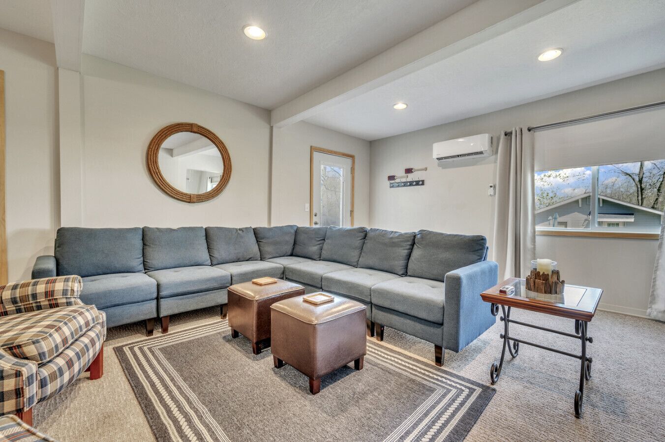 Un salon comprenant un canapé d’angle, un fauteuil à carreaux, deux poufs, une table basse, un miroir, un climatiseur et une fenêtre avec vue.