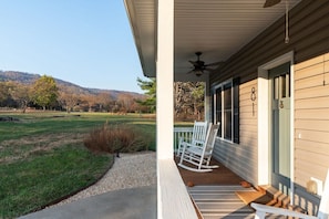 A porch with a ceiling fan, white rocking chairs, and a view of a grassy field and hills.