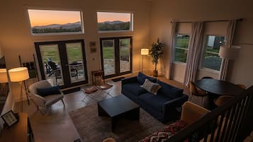 A spacious living room with a large sofa, a coffee table, and a dining table. There are two windows with a view of the outdoors.
