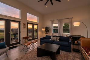 A spacious living room with a dark blue sofa, a coffee table, and a dining table with chairs. Large windows offer views of the outdoors. - Luxury Stay w/ Fenced Yard, Hot Tub, Mountain View (Afton)