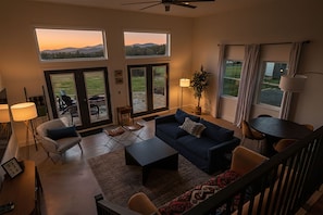 Sala de estar com sofá, cadeiras, mesa de centro, luminária e janelas amplas com vista para o exterior.