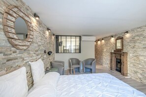 Quarto com parede de tijolos, uma cama grande, duas cadeiras, uma lareira e um espelho.