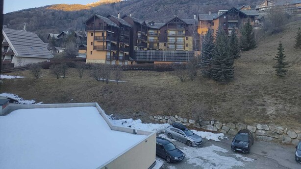 Un edificio de varios pisos con balcones, un estacionamiento con coches y un paisaje montañoso al fondo.