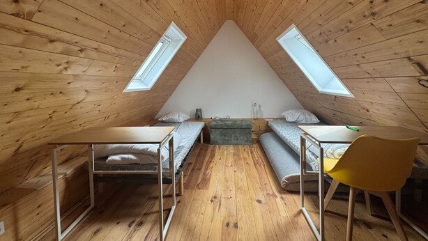 Et soveværelse under skråvæggen med loft, trævægge, to ovenlys, en seng, et skrivebord, en stol og et lille bord.