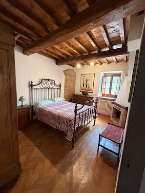 Un dormitorio con techo de madera, una cama con colcha estampada, una mesita de noche, un aparador, un radiador y una ventana.