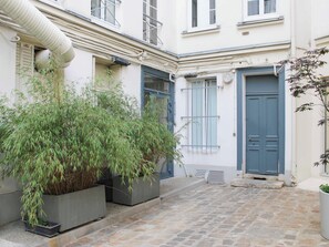 A courtyard with a stone floor, a large potted bamboo plant, and a building with blue doors and shutters. - 96m² flat, overlooking Paris rooftops (Paris)