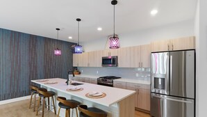 Une cuisine moderne dotée d’un îlot central, de tabourets de bar, d’appareils électroménagers en acier inoxydable et de luminaires suspendus.