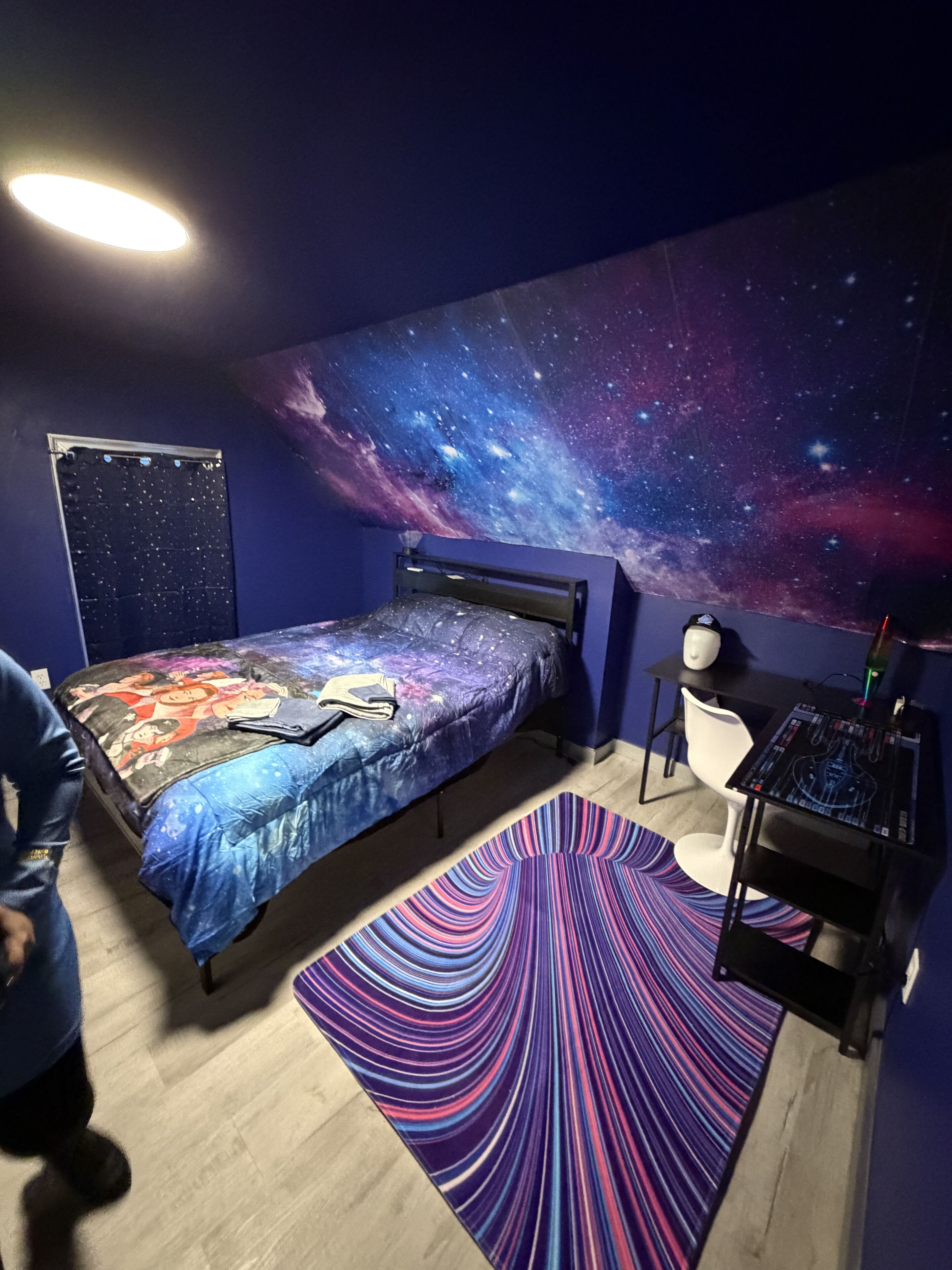 Une chambre avec un lit, un bureau, une chaise, une fresque murale représentant un ciel étoilé et un tapis à motifs.