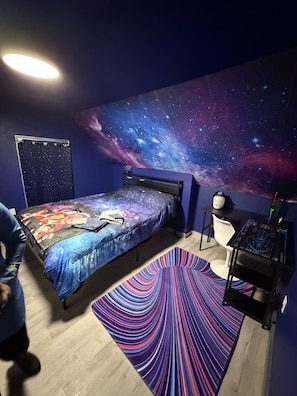 Ein Schlafzimmer mit Bett, Schreibtisch, Stuhl, Wandmalerei mit Sternen und einem gemusterten Teppich.