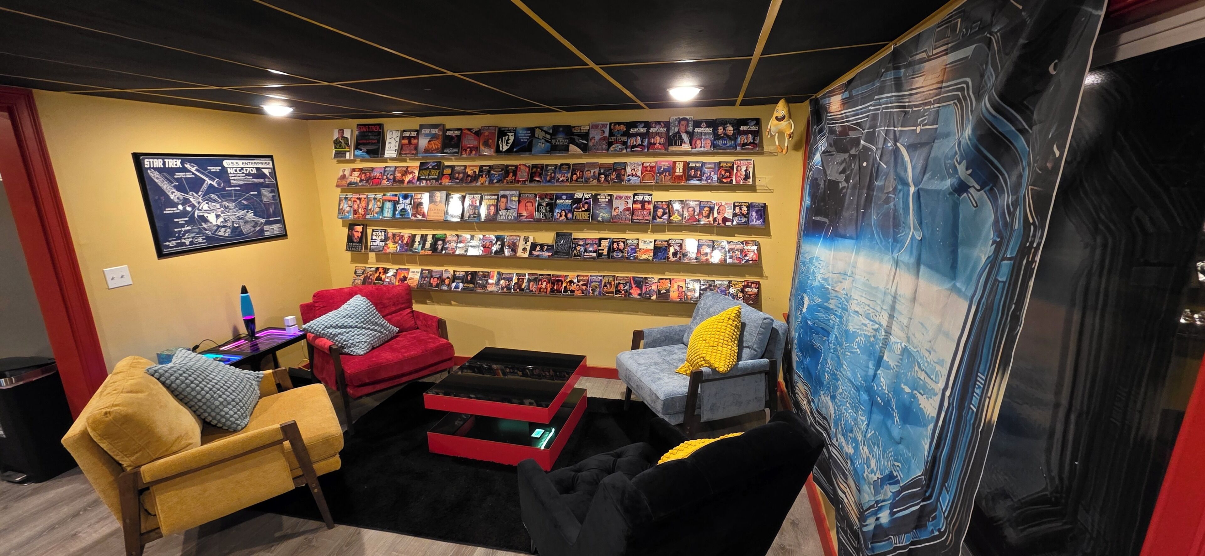 Une pièce comprenant une bibliothèque, un fauteuil rouge, un fauteuil jaune, un pouf noir et une fresque murale bleue.