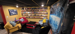 Ein Zimmer mit Bücherregal, einem roten Sessel, einem gelben Sessel, einem schwarzen Hocker und einem blauen Wandbild.