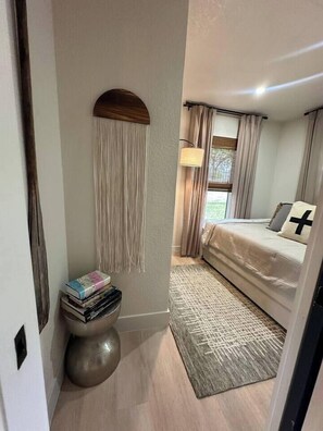 Una camera da letto con un letto, una lampada da comodino, un tavolino rotondo con dei libri, una lampada a parete e una finestra con tende.