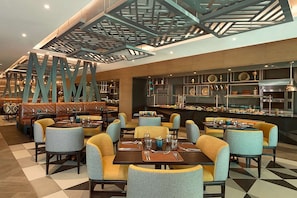 Un restaurant avec de hauts plafonds, des luminaires aux formes géométriques et un mélange de banquettes et de tables.