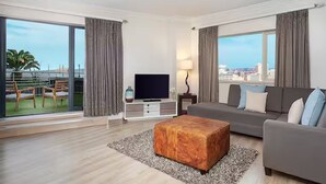 A modern hotel room with a large balcony, a flat-screen TV, a grey sofa, and a wooden ottoman. - Hilton Durban (Durban)