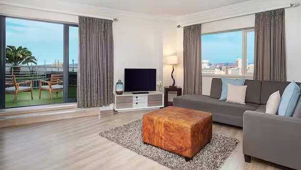 A modern hotel room with a large balcony, a flat-screen TV, a grey sofa, and a wooden ottoman. - Hilton Durban (Durban)